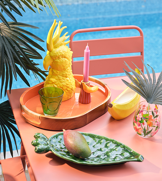Bright outdoor table display with colorful fruit shaped décor, glassware, and candles beside a blue swimming pool.
