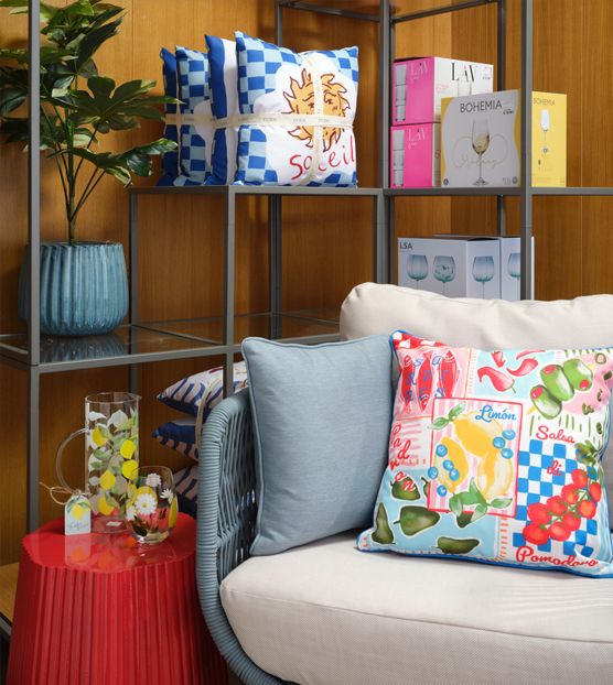 Bright, cosy living setup with a beige sofa, colourful fruit-print cushion, red side table with patterned glassware, and shelving displaying plants, checked cushions, books, and boxed glassware.