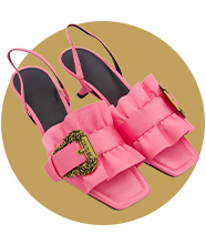 A pair of bright pink slingback heeled sandals with layered ruffle detailing and decorative gold buckles, displayed against a circular gold backdrop.