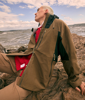 A model sitting on a rock by the seaside wearing a tonal waterproof jacket 