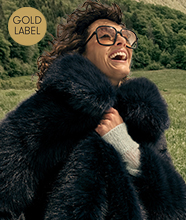 Woman outdoors wearing oversized sunglasses and a dark faux-fur coat. Gold circular sign in the corner reads “GOLD LABEL”.