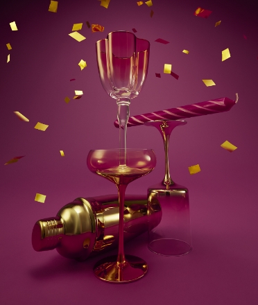 Two stacked coupe glasses with a gold toned cocktail shaker behind. Beside this, a roll of gift wrap lays on top of a champagne glass.