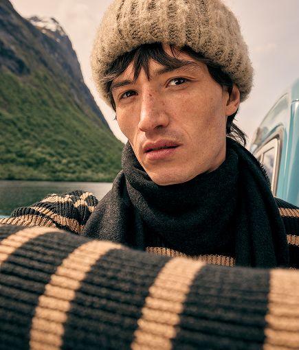 Man wearing a striped knit jumper, scarf and wool hat beside a mountain landscape.