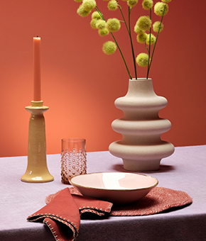 A table with a vase of yellow flowers, candle holder, glass, bowl and folded napkins on a tablecloth.