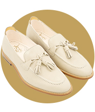 A pair of cream leather loafers with tassel details and stitched trim, displayed at an angle against a circular gold background.