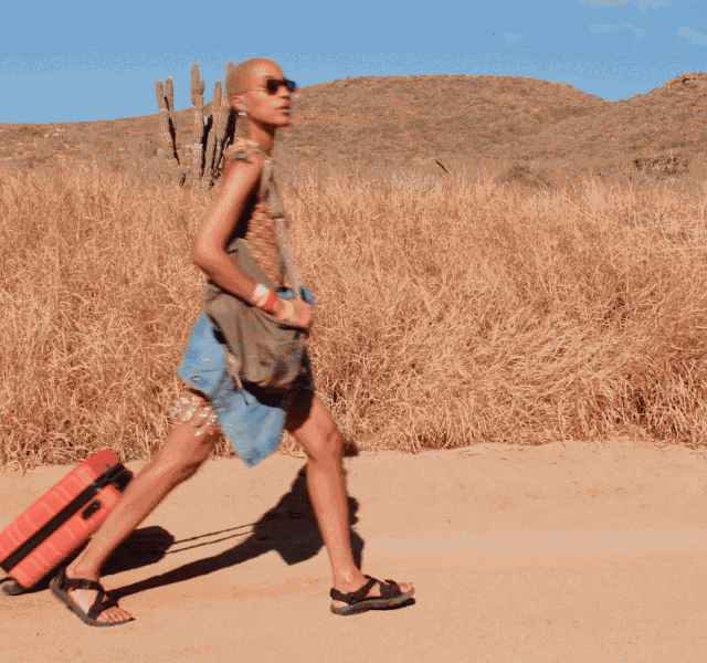A model wearing a fringed mini dress and dark shades wheeling a cabin-sized suitcase. 