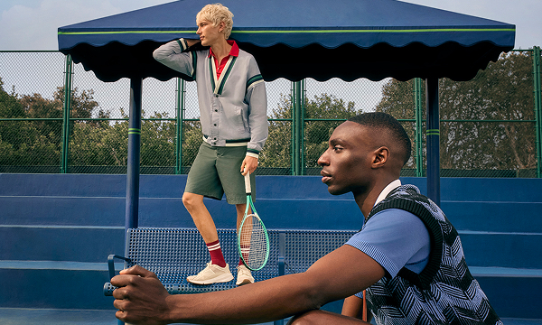 Two male models at a tennis court holding tennis rackets. 				