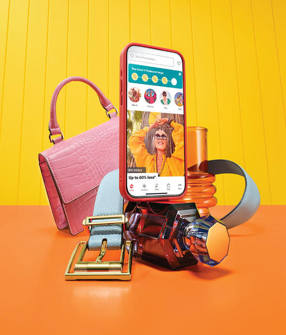 A phone showing the TK Maxx app surrounded by a handbag, belt and accessories against a yellow background.