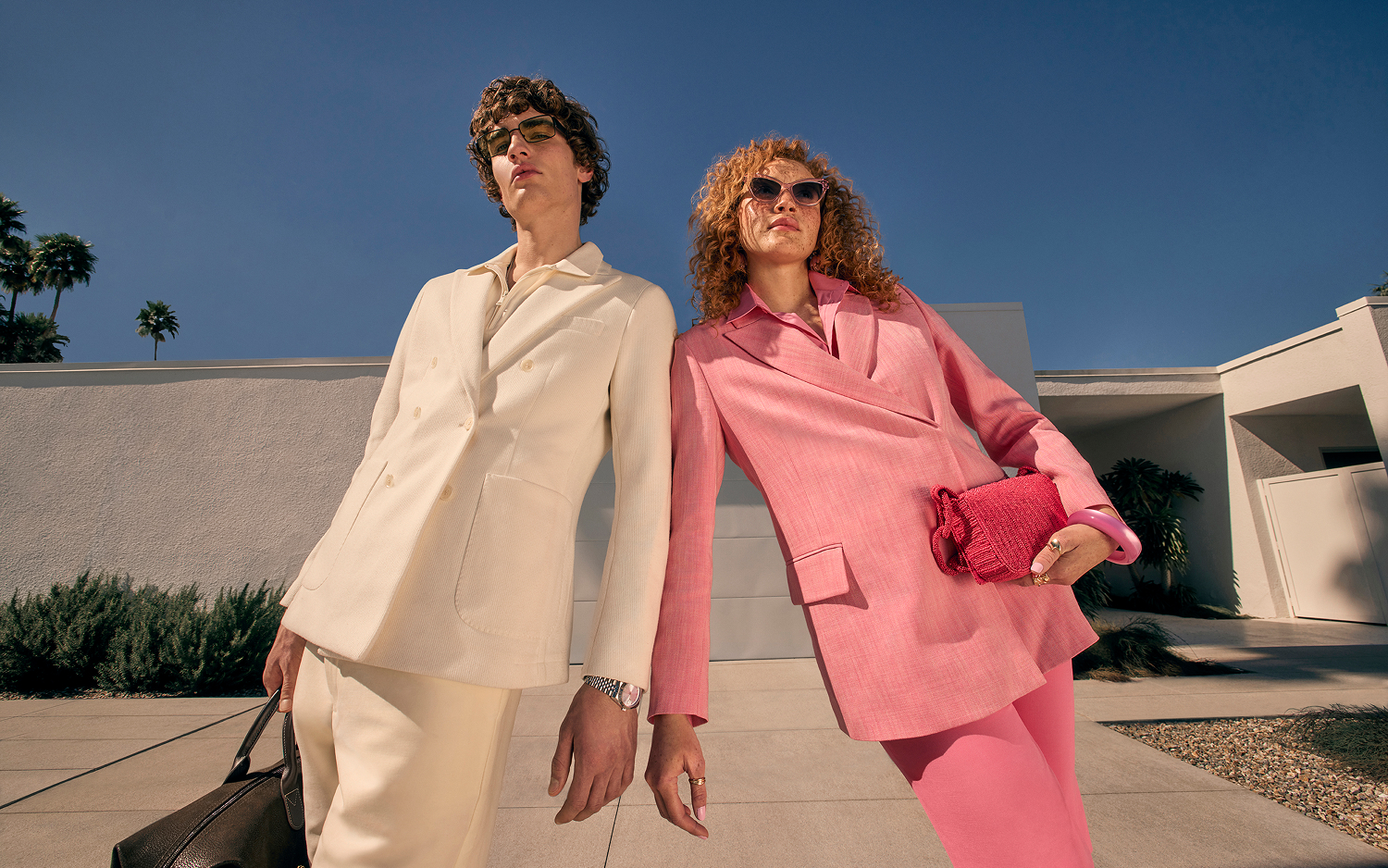Two people dressed in suits, one in cream and one in pink, wearing sunglasses and carrying bags. 