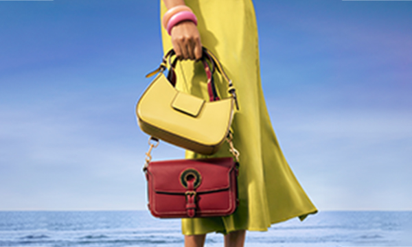 Woman stands on a grassy field in front of the sea. She holds two handbags: one red, one green. She is wearing a green dress and bangles.