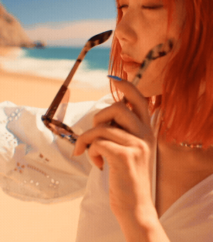 A woman wearing a white summer dress at the beach puts on a pair of tortoiseshell sunglasses.