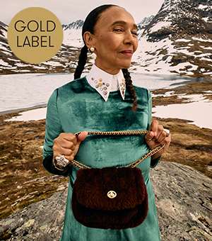 A female model wearing a velvet green dress over a crisp white shirt, holding a luxury leather shoulder bag. 