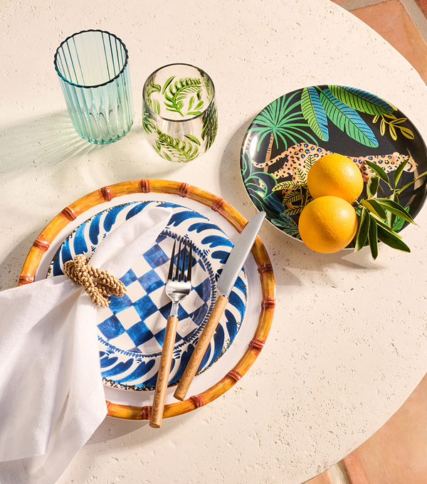 A tabletop place setting with patterned plates, wooden-handled cutlery, two glasses, and a bowl with lemons and greenery on a light surface.