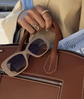 A model wearing gold rings holds a tan bag with beige sunglasses clipped onto the handle.