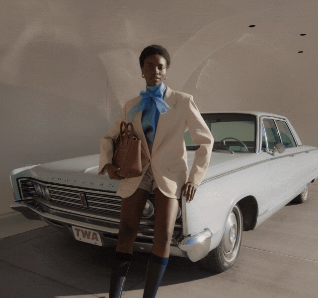 A model standing next to a vintage car wearing shorts, a neutral blazer and bold blue blouse.