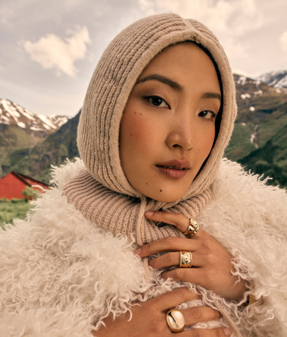 Woman wearing a knitted beige hood and a textured cream coat, with mountains in the background.