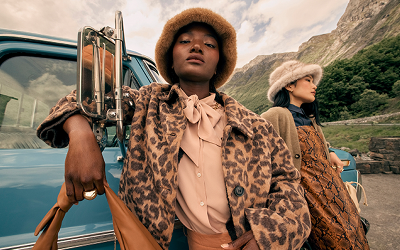 Two models in the countryside wearing faux fur hats and bold, animal print coats.