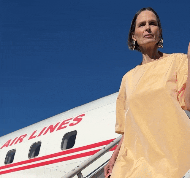 A model in a yellow shift dress stands in front of a plane.