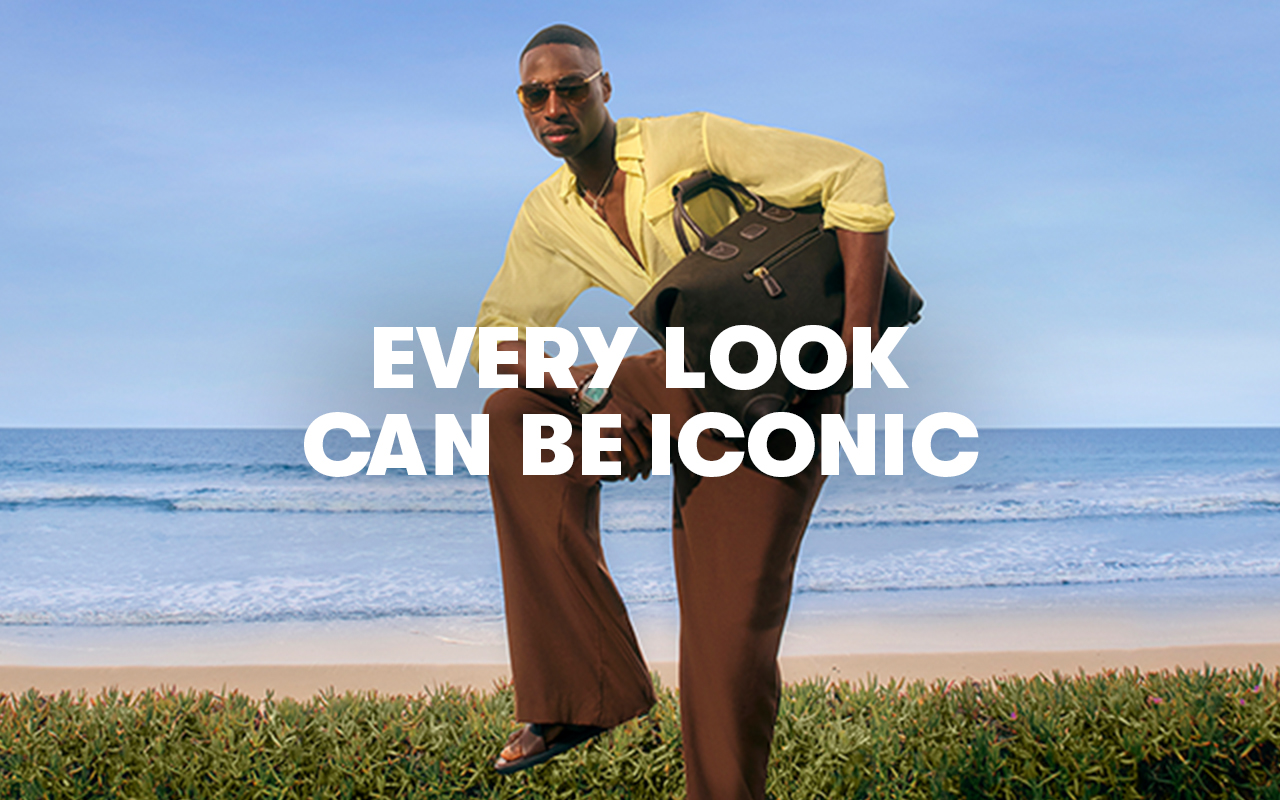 A man poses on some grass in front of a beach. He carries a big leather bag and wears brown trousers with a yellow shirt.