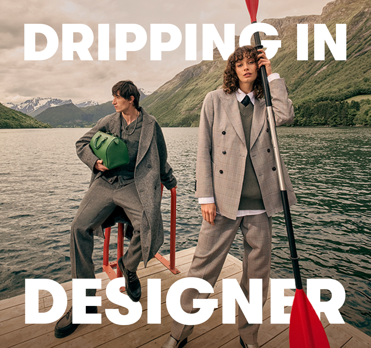 Two people in tailored outfits stand on a lakeside dock with mountains behind them. Text reads: “DRIPPING IN DESIGNER”.