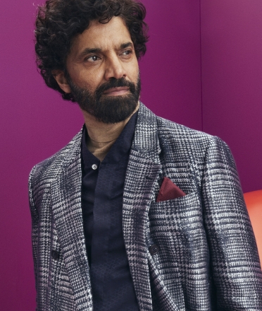 A bearded man wears a navy shirt with a dark blue plaid blazer and purple pocket square.