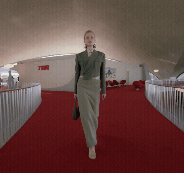 Woman walking down a red-carpeted hallway in a fitted grey jacket and long grey skirt, carrying a bag. She has a slick bob and dangly earrings.				