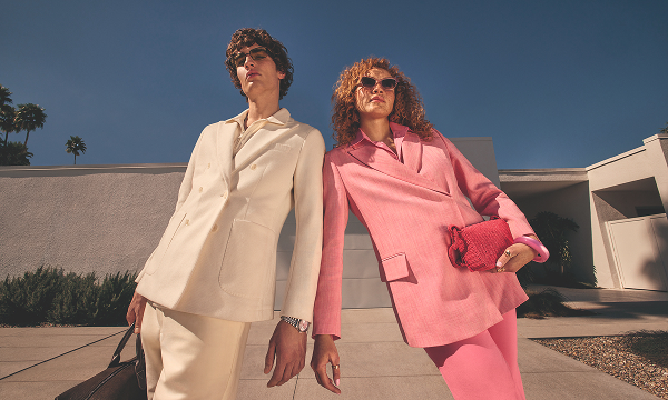 Two people in tailored cream and pink suits lean on each other outdoors, posing in front of a modern concrete building under a clear blue sky.