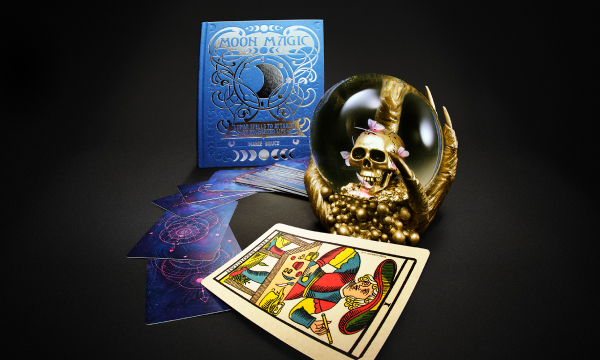 Crystal ball with a skull inside, tarot cards and a moon-themed book on a dark tabletop.
