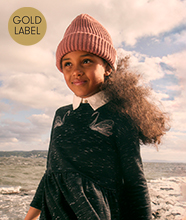 Child wearing a pink knit beanie and dark dress with a white collar stands by the seaside. Gold circular sign in the corner reads “GOLD LABEL”.