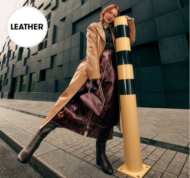 A female model wearing a sleek leather coat, a cosy knit and patent leather midi skirt. 