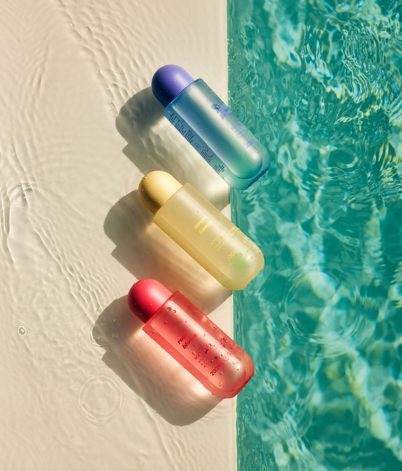 Three colourful bottles rest on the edge of a turquoise pool. The bottles are blue, yellow and red, and cast soft shadows against the water.