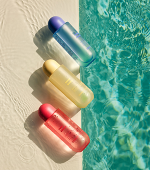 Three colourful skincare bottles in blue, yellow and red lie on a sunlit pool edge, casting soft shadows beside clear rippling water.