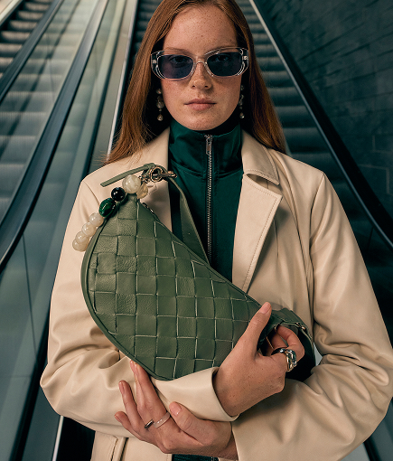 Woman wearing a light-coloured jacket and sunglasses, holding a green bag on an escalator.