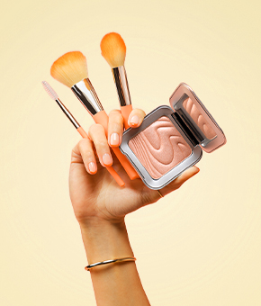 A hand holds makeup brushes and an open highlighting powder compact against a soft yellow background.