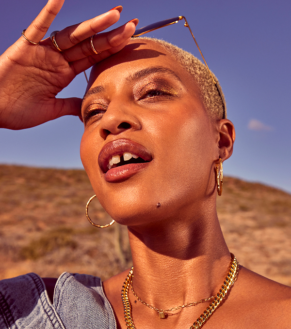 A woman raising a hand to shield her eyes from the sun, wearing gold jewellery and a denim top, with a blue sky and dry landscape in the background.