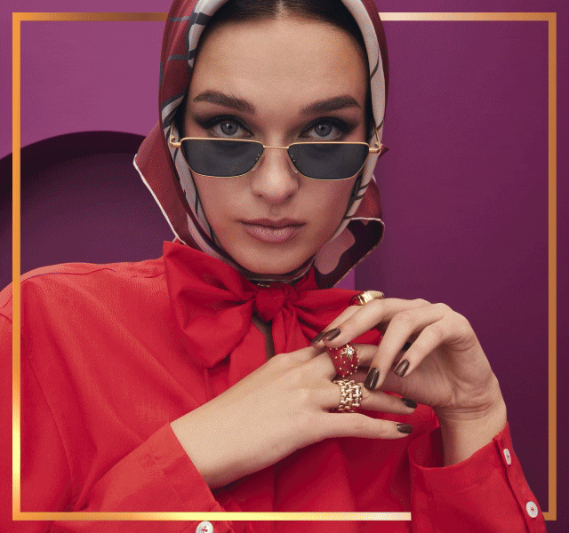A woman wearing a scarf, sunglasses and red blouse, touching the rings she's also wearing.