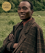 Man standing in a grassy field wearing a checked coat over a shirt and tie. Gold circular sign in the corner reads “GOLD LABEL”.