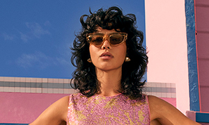 A person in a sleeveless sparkly pink top stands outdoors, with sunglasses on, in front of a pink and blue building under a clear sky.