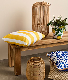 A wooden bench with striped and patterned cushions, a wicker lantern and a potted plant in a lemon pot.