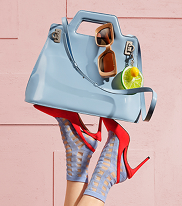 Blue bag with sunglasses and a bag charm balanced on feet wearing red high heels and blue socks.