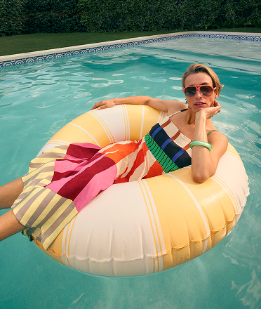 A female model on a pool float wearing a bright dress and statement sunnies.
