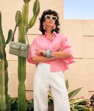 Person wearing a pink blouse, white trousers, sunglasses and bracelets, standing cross-armed outdoors beside a cactus, which has a bag hanging from it.